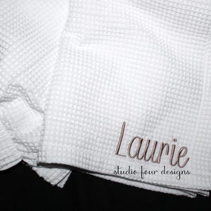 May include: A white waffle-weave towel featuring the embroidered name Laurie in a cursive brown script, with the text studio four designs printed in a smaller black font directly underneath the name on the fabric surface.
