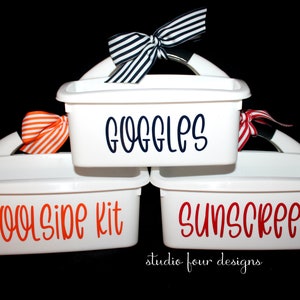May include: Three white plastic storage bins with handles and labels. The top bin is labeled "Goggles" in navy blue lettering. The middle bin is labeled "Poolside Kit" in orange lettering. The bottom bin is labeled "Sunscreen" in red lettering.