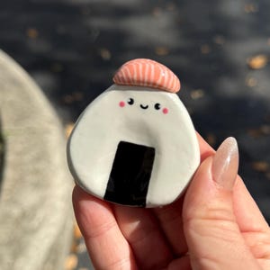 Può includere: Un onigiri in ceramica bianca con un accento di alghe nere e una faccia sorridente. L'onigiri è sormontato da un cappello rosa. L'immagine è di un oggetto decorativo fatto a mano.