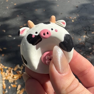 May include: A ceramic cow figurine with a white base, black spots, and a pink nose. The cow has small, curved horns and black ears. The figurine is round and appears to be hand-painted.