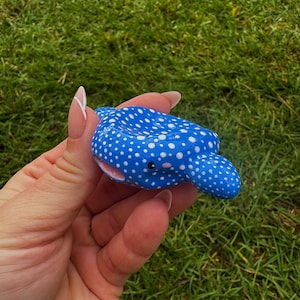May include: A blue and white spotted clay sculpture of a manta ray. The sculpture has a rounded body, small fins, and a smiling face with pink accents. The sculpture is held in a hand against a background of green grass.