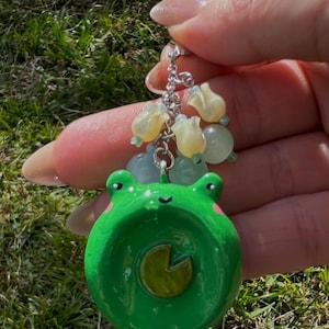 Puede incluir: Un colgante verde con forma de rana con un diseño de nenúfar, mejillas rosas y una cara sonriente. Está unido a una cadena plateada con cuentas amarillas y verdes, creando un accesorio caprichoso.
