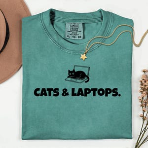 Peut inclure: T-shirt de couleur sarcelle avec le texte "CATS & LAPTOPS." Un chat noir est représenté sur un ordinateur portable. Un collier doré avec un pendentif en forme d'étoile est sur le t-shirt. Un chapeau marron est dans le coin supérieur gauche.