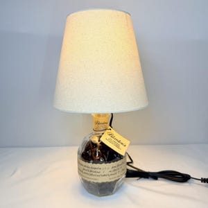 May include: A table lamp featuring a clear glass Blanton's bourbon bottle base filled with dark glass stones. The lamp has a tapered, off-white fabric lampshade and a black electrical cord. A small tan tag with the Blanton's logo is attached.