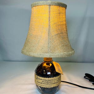 May include: A unique table lamp crafted from a repurposed bourbon whiskey bottle. The lamp features a burlap shade and a warm, inviting glow. The bottle retains its original label and a small tag. A black power cord extends from the base.