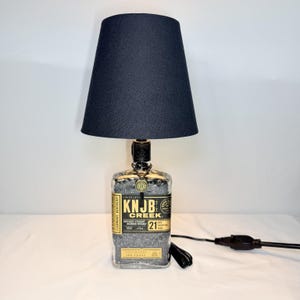May include: A table lamp featuring a clear glass Knob Creek Kentucky Straight Bourbon Whiskey bottle base filled with small dark stones. The bottle has a black label with white text and a black fabric tapered lampshade with a black power cord.