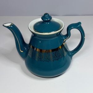 May include: A deep teal ceramic teapot featuring a matching lid with a knob handle. The body is decorated with a wide, horizontal band of intricate gold filigree patterns. The spout and handle are accented with thin gold trim along the edges.
