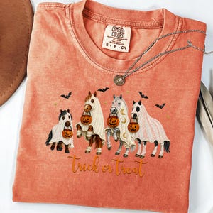 Op de afbeelding: Een koraalkleurig T-shirt met korte mouwen en een afbeelding van vier paarden verkleed als spoken, elk met een pompoenemmer. De tekst "trick or treat" staat onder de paarden gedrukt, omringd door kleine zwarte vleermuizen in het ontwerp.