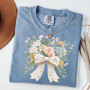 Puede incluir: Una camiseta azul Comfort Colors con un diseño de ramo floral y un lazo grande. El ramo incluye flores rosas, blancas, amarillas y azules. Un collar dorado con un colgante está drapeado sobre la camisa.