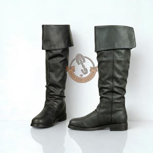 May include: A pair of knee-high black boots with a folded cuff at the top. The boots feature a smooth finish and a low heel. A circular logo with the text IRON STBP LEATHER and a chain link graphic is positioned between the two boots.