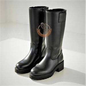 Puede incluir: Un par de botas de cuero negro a media pantorrilla con punta redondeada, suela gruesa de tacos y cierre de cremallera lateral. Un parche circular marrón con el texto IRON STEP LEATHER y un gráfico de huella de bota es visible en el lateral.