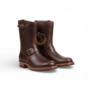May include: A pair of brown leather boots with a mid-calf height. Each boot features a buckle closure and a wooden sole. The boots have the text "IRON STEP LEATHER" on the side.