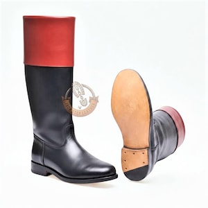 Puede incluir: Un par de botas altas de cuero con una sección inferior negra y un puño rojo en contraste en la parte superior. Una bota está de pie, mientras que la otra está colocada para mostrar la suela de cuero beige y un tacón pequeño. Se ve el logotipo Iron Step Leather.