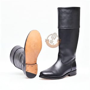 Pode incluir: Um par de botas de cano alto em couro preto com acabamento liso e uma dobra larga no topo. Uma bota está em pé, enquanto a outra está posicionada para mostrar a sola de couro bege e um pequeno salto com pregos visíveis na base.