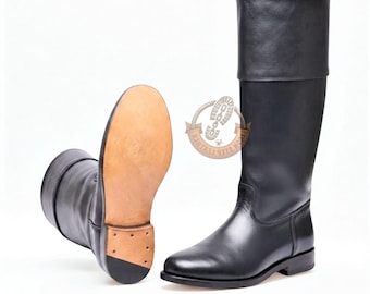 Puede incluir: Un par de botas altas de cuero negro con acabado liso y una solapa ancha en la parte superior. Una bota está de pie, mientras que la otra está colocada para mostrar la suela de cuero color canela y un pequeño tacón con clavos visibles.