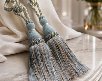 Könnte beinhalten: Zwei dekorative Vorhanghalter mit hellblauen und goldenen gedrehten Seilen und kunstvollen Knoten. Jeder Halter endet in einer großen, vollen Quaste aus feinen, mehrfarbigen hellblauen und goldenen Fäden, verziert mit einem gewickelten Kragen.