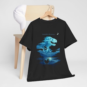Puede incluir: Camiseta negra con una ilustración azul de un Tyrannosaurus Rex en un paisaje con un lago, una gallina y una luna creciente. El texto "Remember who you are" está encima del dinosaurio.