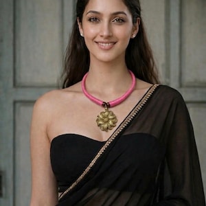 Könnte beinhalten: Eine Frau trägt ein schwarzes trägerloses Oberteil und einen transparenten schwarzen Sari mit goldenem Besatz. Um ihren Hals trägt sie eine rosa Halskette mit einem goldenen Blumenanhänger. Sie lächelt und trägt kleine Ohrringe.