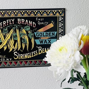 Op de afbeelding: Een zwart vintage bord met de tekst "Butterfly Brand" en "Golden Wax Stringless Bean". Het bord heeft illustraties van bonen en een vlinder. Een boeket bloemen, waaronder tulpen en chrysanten, staat op de voorgrond.