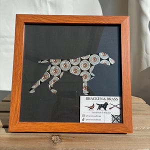 May include: A framed wall art piece featuring a dog silhouette constructed from silver and copper shotgun shell casings on a black background. The wooden frame is light brown. A small white business card with the text BRACKEN & BRASS is included.