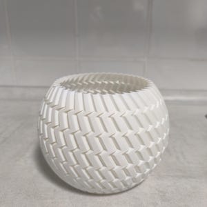 Könnte beinhalten: Eine weiße, kugelförmige Vase mit gewebter Textur. Die Vase hat eine breite Öffnung und einen abgerundeten Körper. Das strukturierte Muster besteht aus sich wiederholenden, abgewinkelten rechteckigen Formen. Die Vase ist wahrscheinlich aus 3D-gedrucktem Material.