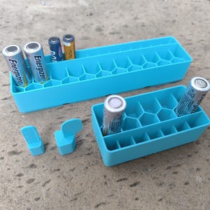 Könnte beinhalten: Zwei hellblaue Batterie-Organizer aus Kunststoff mit einem wabenförmigen Innengitter. Das größere rechteckige Fach enthält mehrere AA-Batterien, während das kleinere Fach eine AA-Batterie enthält. Zwei kleine blaue Kunststofflaschen liegen separat.