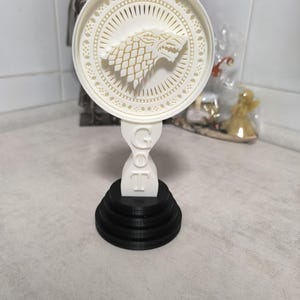 K&ouml;nnte beinhalten: Wei&szlig;-schwarzes Deko-Objekt mit einem Wolfskopf-Emblem in einem kreisf&ouml;rmigen, ausgeschnittenen Design. Der Sockel ist abgestuft und schwarz, mit den Buchstaben "GOT" sichtbar. Wahrscheinlich ein dekorativer Artikel.