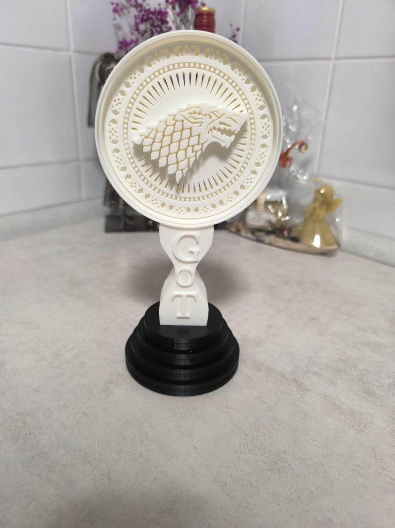 K&ouml;nnte beinhalten: Wei&szlig;-schwarzes Deko-Objekt mit einem Wolfskopf-Emblem in einem kreisf&ouml;rmigen, ausgeschnittenen Design. Der Sockel ist abgestuft und schwarz, mit den Buchstaben "GOT" sichtbar. Wahrscheinlich ein dekorativer Artikel.