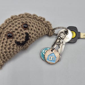May include: A tan crocheted dumpling-shaped keyring with two dark brown plastic eyes and a black stitched smile. A silver metal ring is attached, holding a silver Yale key and two small circular charms, one featuring a light blue house icon.