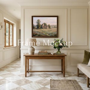 May include: A framed landscape painting hangs above a wooden console table in a well-lit room. The painting depicts a rural scene with a church. A lamp, books, and a vase of flowers are on the table. The room has a neutral color palette with a patterned floor.