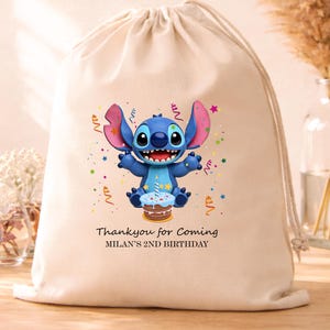 May include: A beige drawstring bag featuring a cartoon character with blue fur, large ears, and a wide smile. The character is holding a cake. Text reads "Thank you for Coming" and "MILAN'S 2ND BIRTHDAY".