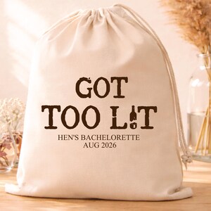 Puede incluir: Una bolsa beige con cordón y la frase "GOT TOO LIT" en marrón. Debajo, se lee "HEN'S BACHELORETTE AUG 2026". La bolsa es de tela suave y tiene cierre de cordón. El fondo incluye elementos decorativos.