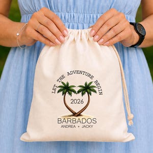 May include: A cream drawstring bag with the text "LET THE ADVENTURE BEGIN" and "2026" printed on it. The bag features a design of two palm trees forming a heart shape, with the word "BARBADOS" and names printed below.