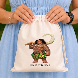 May include: A beige drawstring bag featuring a colorful illustration of the character Maui from the animated film Moana. Maui is depicted holding his magical fish hook. The text "MILO TURNS 3" is printed below the character.