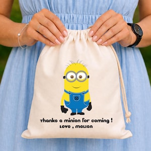 Könnte beinhalten: Beigefarbener Kordelzugbeutel mit einem gelben Minion-Charakter mit zwei Augen, der blaue Latzhosen trägt. Der Beutel trägt den schwarzen Text "thanks a minion for coming! Love, Melisa".