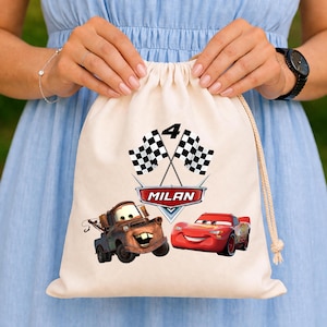 Puede incluir: Una bolsa beige con cordón que presenta el número 4, banderas a cuadros cruzadas y el nombre "MILAN" en una pancarta roja y blanca. La bolsa también muestra imágenes de dibujos animados de Mate y Rayo McQueen de la película Cars.