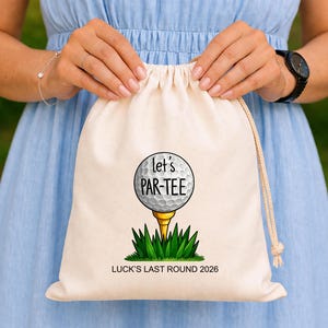 Pode incluir: Uma bolsa bege com cordão com um design de bola de golfe. A bolsa tem o texto "Let's PAR-TEE" e "LUCK'S LAST ROUND 2026". A bola de golfe é branca com texto preto, em um tee dourado e grama verde.