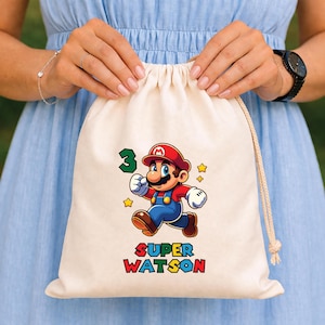 Op de afbeelding: Een beige trekkoordtas met een kleurrijke afbeelding van een stripfiguur met een rode hoed en blauwe overall. Het nummer 3 en de woorden "SUPER WATSON" staan ook op de tas. De tas wordt vastgehouden door een persoon in een blauwe jurk.