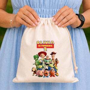 May include: A beige drawstring bag featuring a colorful graphic of characters from the Toy Story movie. The bag has the text "OLIVIA IS TURNING 2" in blue, red, and yellow. The bag is held by a person wearing a blue dress.