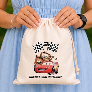 May include: Cream-coloured drawstring bag with a red race car and a brown truck, plus checkered flags and the number 3. The text "RACHEL 3RD BIRTHDAY" is printed below. The bag is held by a person.