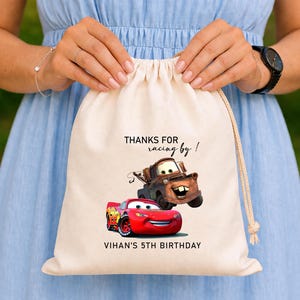 May include: A beige drawstring bag with the text "THANKS FOR racing by!" and "VIHAN'S 5TH BIRTHDAY". The bag displays a cartoon image of a red race car and a brown tow truck.