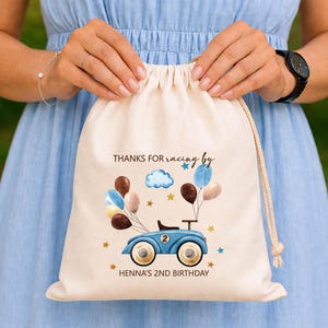 Puede incluir: Una bolsa de algodón color crema con cordón que presenta un gráfico impreso de un coche de carreras de juguete azul con el número 2, rodeado de globos marrones, azules y crema, estrellas y una nube. El texto dice: THANKS FOR racing by HENNA'S 2ND BIRTHDAY.