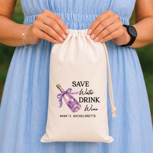 Op de afbeelding: Een crèmekleurige stoffen tas met trekkoord voorzien van een afbeelding van een champagnefles met een paars lint. De tekst luidt SAVE Water DRINK Wine en NANA'S BACHELORETTE in zwarte letters. De tas is gemaakt van lichtgekleurd textielmateriaal.