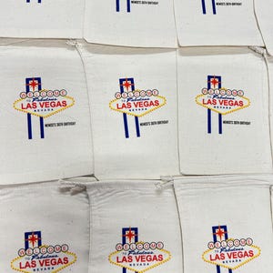 May include: White fabric bags with a printed design of the iconic "Welcome to Fabulous Las Vegas" sign in red, yellow, and blue. The text "MEMOS'S 36TH BIRTHDAY" is printed below the sign. The bags are arranged in a grid.