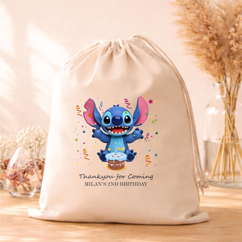 May include: A beige drawstring bag featuring a cartoon character with blue fur, large ears, and a wide smile. The character is holding a cake. Text reads "Thank you for Coming" and "MILAN'S 2ND BIRTHDAY".