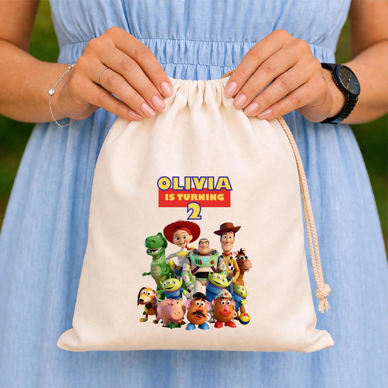 May include: A beige drawstring bag featuring a colorful graphic of characters from the Toy Story movie. The bag has the text "OLIVIA IS TURNING 2" in blue, red, and yellow. The bag is held by a person wearing a blue dress.