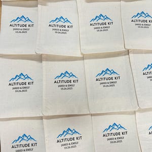 May include: Off-white fabric pouches with a blue mountain graphic and the words "ALTITUDE KIT," "JARED & EMILY," and the date "10.26.2025" printed in black. The pouches are arranged in a grid pattern.
