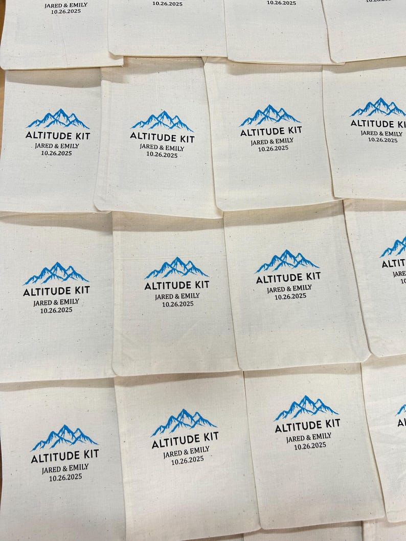 May include: Off-white fabric pouches with a blue mountain graphic and the words "ALTITUDE KIT," "JARED & EMILY," and the date "10.26.2025" printed in black. The pouches are arranged in a grid pattern.