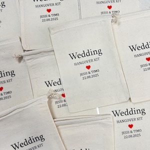 May include: Cream-coloured fabric bags printed with "Wedding HANGOVER KIT" in black, a red heart, and the names "Jessi & Timo" with the date "22.08.2025". Each bag is secured with a drawstring.