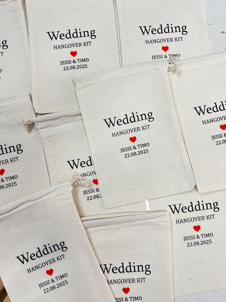 May include: Cream-coloured fabric bags printed with "Wedding HANGOVER KIT" in black, a red heart, and the names "Jessi & Timo" with the date "22.08.2025". Each bag is secured with a drawstring.